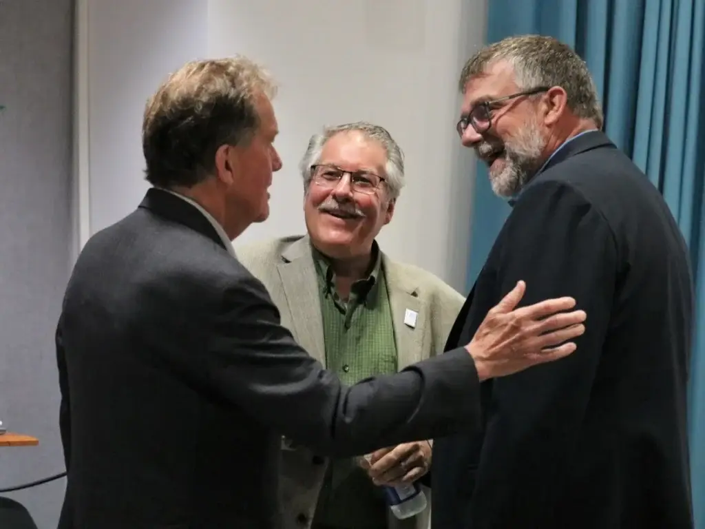 Three men in suits are standing indoors, engaged in conversation. One man gestures with his hand on another's arm. All appear to be smiling. Blue curtain is visible in the background. Liberals Canada Politics